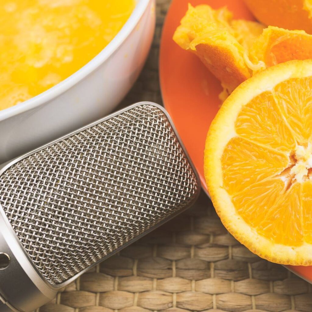 A metal microphone lies next to a sliced orange and a bowl of orange segments on a woven surface
